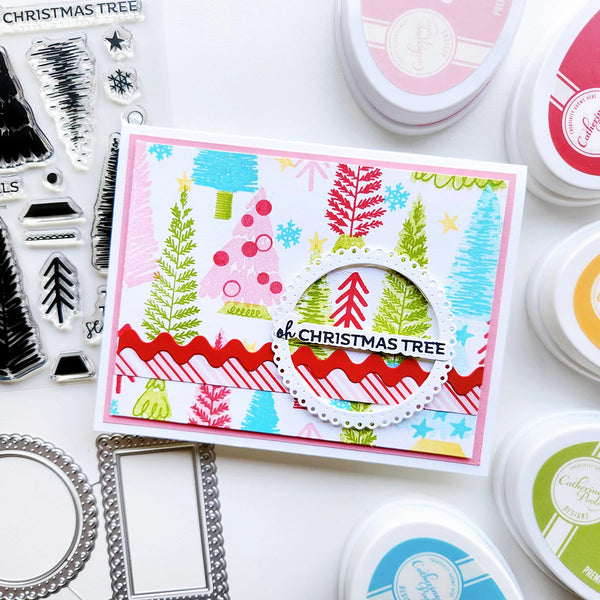 A handmade Christmas card featuring colorful stamped trees, snowflakes, stripes, and a die-cut circle with the text 'of CHRISTMAS TREE', surrounded by crafting tools and ink pads on a white background.