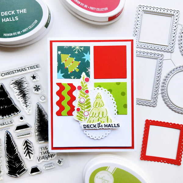 A handmade greeting card featuring a festive Christmas tree design and the words 'DECK THE HALLS,' styled on a white surface with craft supplies including ink pads, clear stamps, and metal frames.