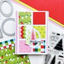A handmade Christmas card with a pink border and a 3x3 grid of colorful holiday patterns, including Christmas lights, stars, stripes, and plaid, with a layered tree and 'DECK THE HALLS' stamp. Surrounding the card are crafting tools including metal die-cut frames, a stamp block, and patterned paper.