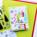A handmade layered Christmas card with colorful tree cutouts, snowflakes, stars, and the phrase 'Tis the season' on a white card base, styled on bright yellow paper with a clear stamp sheet and patterned paper visible at the edges.