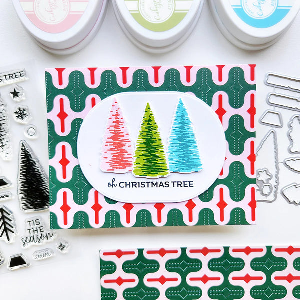 A handmade Christmas card featuring three textured Christmas trees in red, green, and blue on a white oval, set against a green and red patterned card. The card is displayed on a white background with crafting supplies, including stamping tools and ink pads.