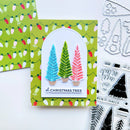 A handmade Christmas card featuring three textured, colorful Christmas trees in blue, green, and pink, set against a green patterned background with holiday lights. The card is displayed alongside crafting tools and a matching patterned paper sheet.