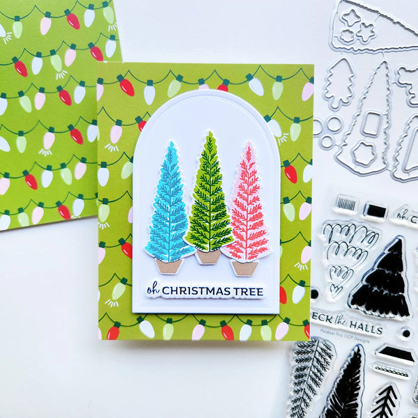 A handmade Christmas card featuring three textured, colorful Christmas trees in blue, green, and pink, set against a green patterned background with holiday lights. The card is displayed alongside crafting tools and a matching patterned paper sheet.