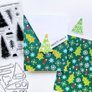 A flat lay of Christmas-themed crafting supplies on a white background, including patterned paper with green trees and snowflakes, a small die-cut tree sticker, clear stamp sheets with tree designs and holiday text, and metal cutting dies.