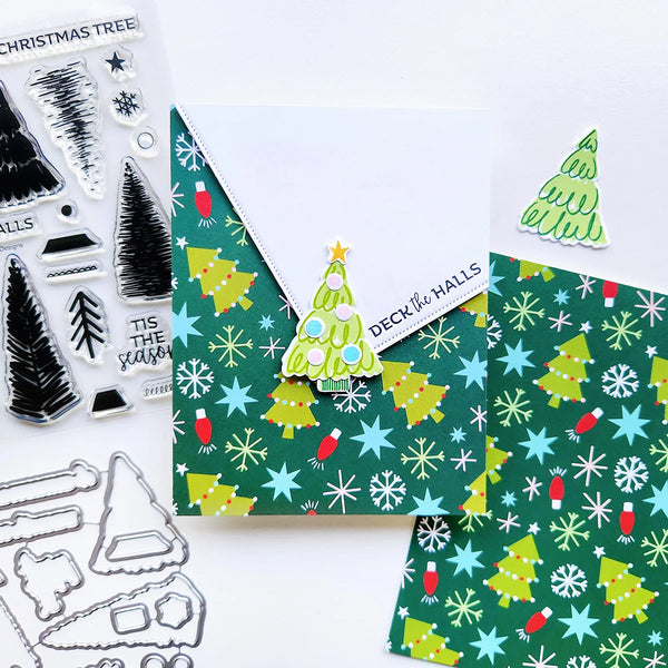 A flat lay of Christmas-themed crafting supplies on a white background, including patterned paper with green trees and snowflakes, a small die-cut tree sticker, clear stamp sheets with tree designs and holiday text, and metal cutting dies.