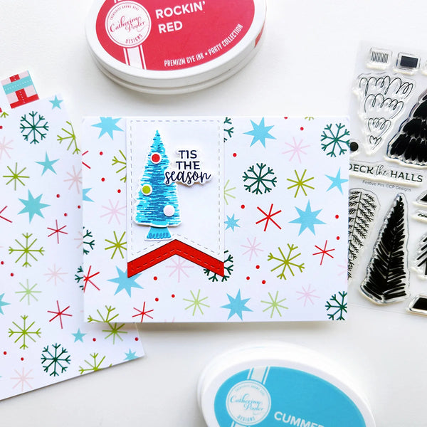 A handmade Christmas greeting card featuring a blue glittery tree and the text 'Tis the season' surrounded by colorful snowflakes and stars, displayed with crafting supplies including Dye Ink pads and clear stamps on a white background.