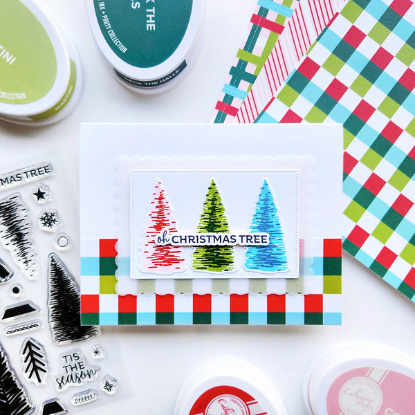 A flat-lay of a handmade Christmas card featuring three colorful Christmas trees, surrounded by crafting supplies including ink pads, patterned paper, and clear stamps on a white background.