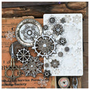 A flat-lay of steampunk-themed metal craft embellishments, featuring silver and pewter snowflakes, cogs, and gears, arranged on vintage newspaper and wood with a white silicone mold sheet on the right.