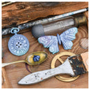 A styled flat-lay of vintage-inspired craft or jewelry-making supplies on a wooden surface, including a blue pocket-watch-shaped charm, a butterfly-shaped embellishment, a spoon with blue beads, a glass vial, a compass, and a paintbrush.