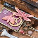 A styled flat-lay of two pink dragonfly and moth-shaped decorative pieces arranged on a purple card over a wooden surface with vintage-style tools and props.