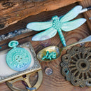 A styled flat-lay of vintage-inspired metal decor on a rustic wooden surface, featuring a turquoise dragonfly ornament, a matching turquoise pocket watch, a bronze-toned spoon, and an ornate bronze medallion.