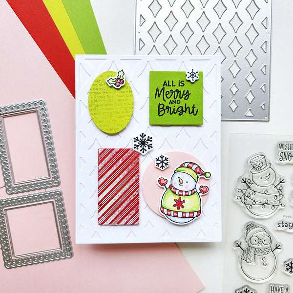 A handmade Christmas greeting card displayed on a styled flat-lay background with crafting tools, including metal die-cut frames, a laser-cut pattern sheet, and snowman-themed clear stamps.