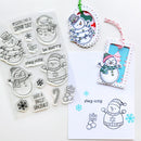 A sheet of clear winter-themed stamp designs with snowman illustrations and phrases like 'stay cozy' and 'HAVE A JOLLY HOLIDAY,' shown beside stamped examples on white cards and hanging tags.