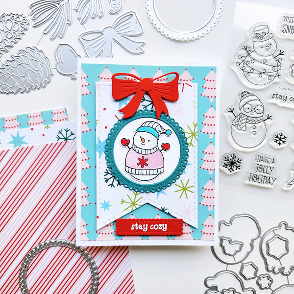 A handmade Christmas greeting card featuring a snowman illustration, a red bow, and a red banner that says 'stay cozy', surrounded by crafting tools, paper, and stamped designs on a white background.