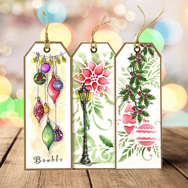 Three decorative hanging gift tags with Christmas illustrations are displayed on a wooden surface against a soft blurred holiday bokeh background. The left tag shows colorful hanging ornaments with the word 'Boule' at the bottom.