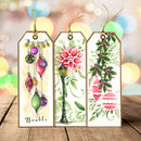 Three decorative hanging gift tags with Christmas illustrations are displayed on a wooden surface against a soft blurred holiday bokeh background. The left tag shows colorful hanging ornaments with the word 'Boule' at the bottom.