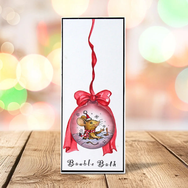 A vertical rectangular card or ornament-style print with a white background, featuring a red hanging ribbon and bow around a round ornament illustration of a mouse in the snow. The card is photographed standing on a wooden surface with colorful blurred bokeh lights in the background.