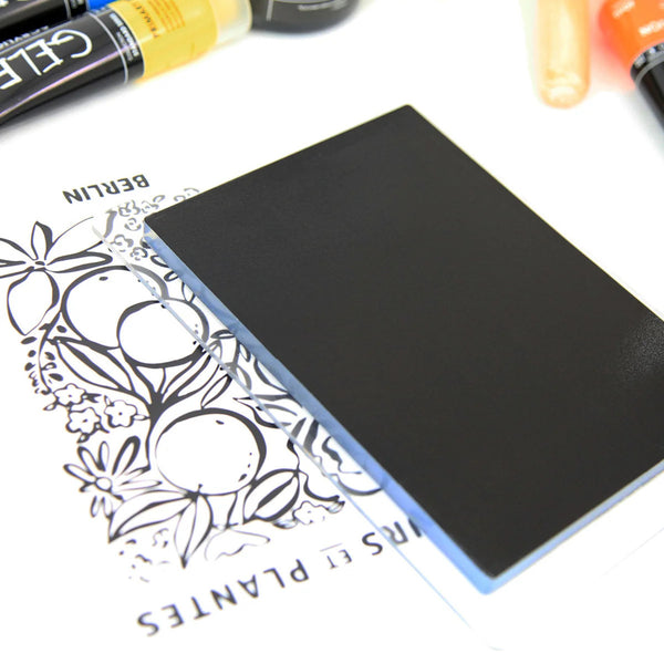 A black rectangular card or mat with a matte finish, shown resting on top of a white printed sheet with botanical line art. Partial colorful pens or markers are visible along the top edge of the frame.