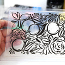 A hand holds a clear rectangular plastic sheet featuring a black line art design of citrus fruits, flowers, and leaves. The plastic has a subtle iridescent sheen, and the background is softly blurred.
