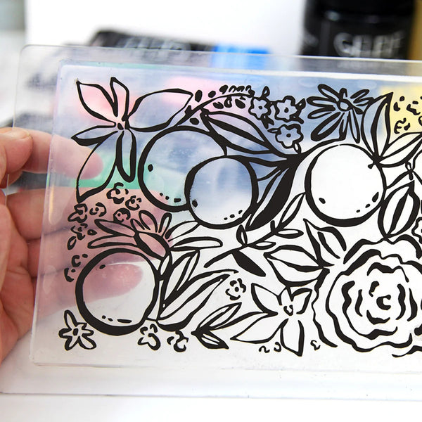 A hand holds a clear rectangular plastic sheet featuring a black line art design of citrus fruits, flowers, and leaves. The plastic has a subtle iridescent sheen, and the background is softly blurred.
