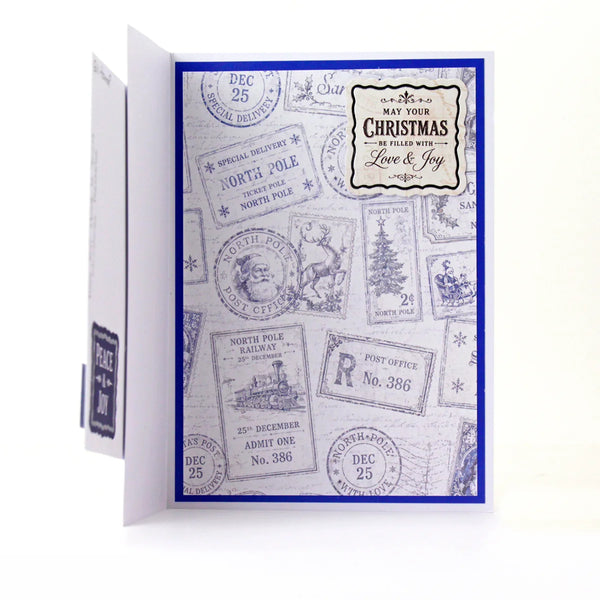 A handmade Christmas greeting card with a blue border and a collage of vintage postage stamp designs in blue ink on a white cardstock base. The front features a central label that reads 'MAY YOUR CHRISTMAS BE FILLED WITH Love & Joy'.