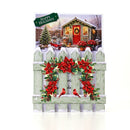 A square Christmas greeting card featuring a snowy winter cottage scene with a green fence, red poinsettias, pinecones, two red birds, and a green 'Happy Holidays' banner at the top.