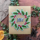 A handmade kraft paper greeting card featuring a die-cut holly wreath with red berries and a central silver 'JOY' star embellishment, styled on a rustic brown surface with green leaves and fabric.