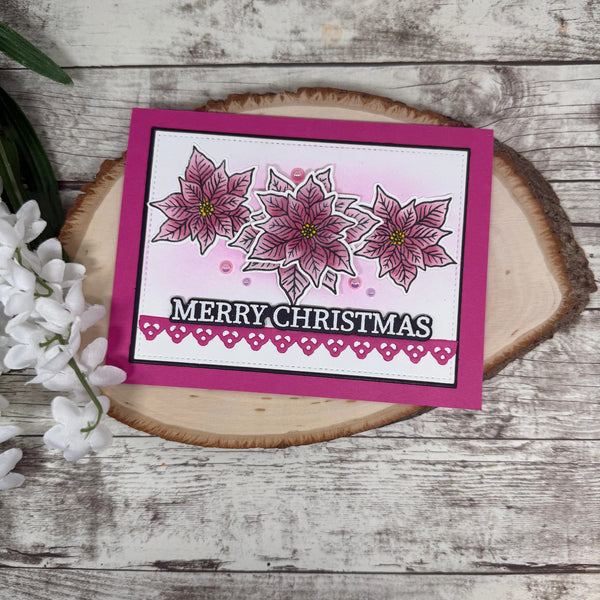 A handmade Christmas greeting card with three pink poinsettia illustrations and the text 'MERRY CHRISTMAS' on a white background, set on a wooden slice coaster with white flowers and green leaves.