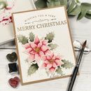 A handmade Christmas greeting card with watercolor poinsettias and green leaves, styled on a white wooden surface with a paintbrush, ink blocks, and eucalyptus leaves.