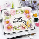 A handmade greeting card with a watercolor floral design and a butterfly, placed on a white surface with a paintbrush, paint palettes, and flowers around it.
