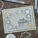 A handmade sympathy card with layered floral designs in white, gray, and gold, featuring the text 'With Sympathy' in black script. The card is displayed on a wooden surface alongside crafting tools, including markers and a stamping plate.