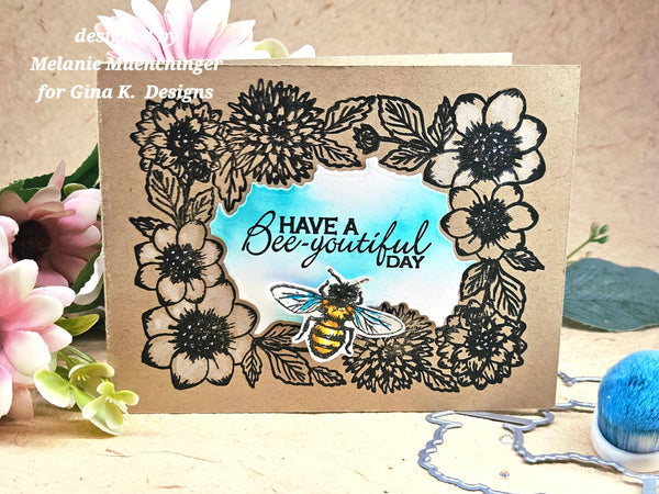 A handmade greeting card with a kraft paper background, featuring a black ink floral border, a blue watercolor center, a bee illustration, and the text 'HAVE A Bee-youtiful DAY'. The card is styled with pink flowers and a blue pom-pom on a wooden surface.
