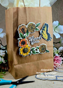 A brown kraft paper gift bag decorated with a floral and butterfly sticker design that reads 'Thank You,' shown with scissors and flowers around it on a tabletop.