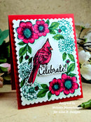 A handmade greeting card featuring a red cardinal, pink and teal flowers, green leaves, and the word 'Celebrate!' on a scalloped white border over a red card base. The card is photographed standing upright on a textured beige surface with blurred flowers in the background.