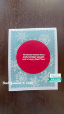 A handmade holiday greeting card on a dark wood background. The card features a white base with a scalloped light blue border decorated with white snowflakes, a large red circle in the center containing the text 'Warmest wishes for a joyful holiday season and a happy New Year.