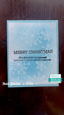 A handmade Christmas greeting card with a light blue and white watercolor gradient background, embossed snowflakes, and the text 'MERRY CHRISTMAS' with a message below. The card is photographed on a dark wooden surface, with a 'gina k.