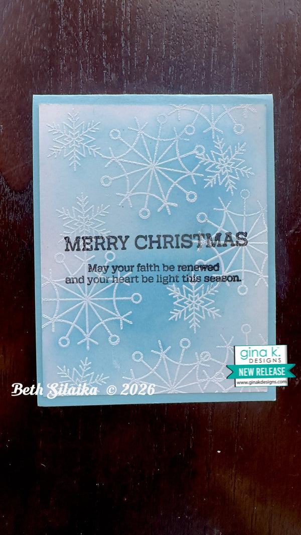 A handmade Christmas greeting card with a light blue and white watercolor gradient background, embossed snowflakes, and the text 'MERRY CHRISTMAS' with a message below. The card is photographed on a dark wooden surface, with a 'gina k.