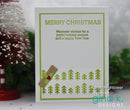 A handmade Christmas greeting card with green text reading 'MERRY CHRISTMAS' and a holiday message, decorated with green pine tree stamps and a small brown envelope with a red holly accent. The card is displayed on a white surface with blurred festive decorations in the background.
