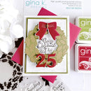 A handmade Christmas greeting card featuring a gold wreath with a red bow and the words 'Merry Christmas to You' above large red '25' numerals. The card is styled with pink paper, stamp pads, and white flowers on a white background with 'gina k.