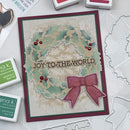 A handmade Christmas greeting card with a layered holly wreath design, red berries, and a pink ribbon bow, placed on a white surface with stamp pads and die cutters around it. The card reads 'JOY TO THE WORLD'.