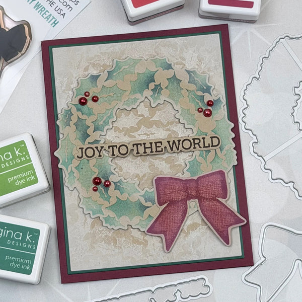 A handmade Christmas greeting card with a layered holly wreath design, red berries, and a pink ribbon bow, placed on a white surface with stamp pads and die cutters around it. The card reads 'JOY TO THE WORLD'.