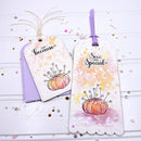 Two decorative paper gift tags with scalloped edges, watercolor autumn pumpkin designs, and printed phrases 'Just Because' and 'Sew Your Special,' displayed on a white wood background with scattered confetti. A lavender ribbon and backing card are visible.