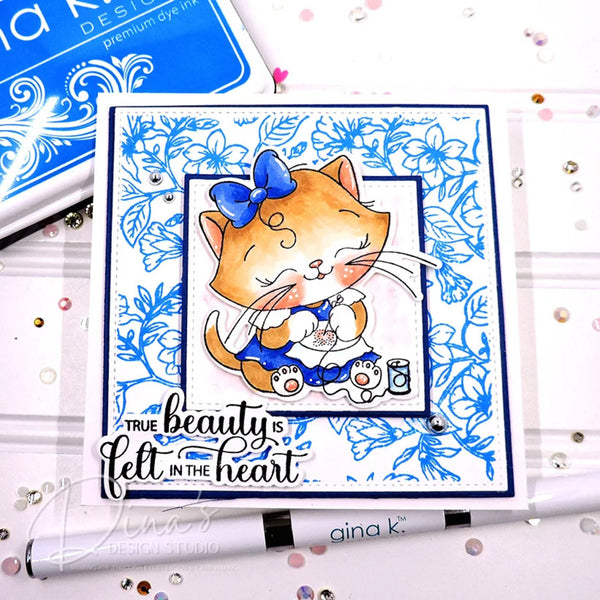 A handmade greeting card featuring a cute orange tabby cat in a blue bow and blue outfit, surrounded by blue floral patterns. The card includes the text 'TRUE beauty is felt in the heart' and is displayed on a white surface with craft tools and rhinestones.