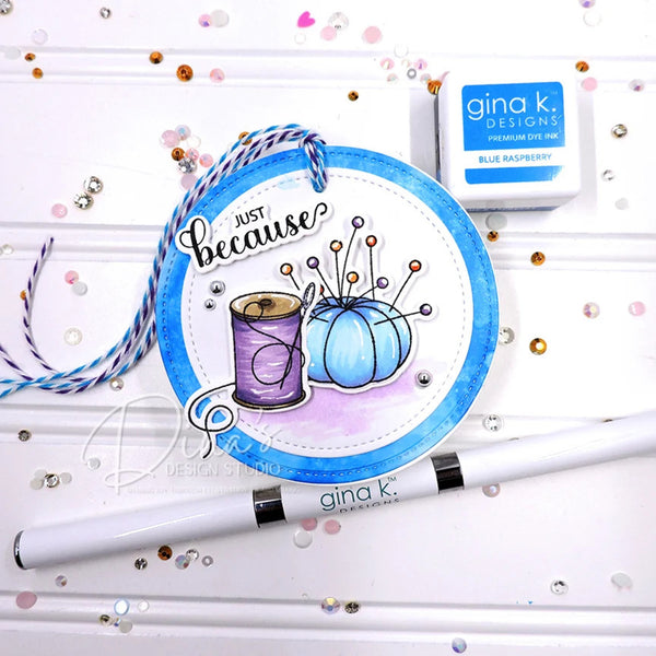 A handmade greeting card or craft paper tag with a blue circular border, the phrase 'Just because' in script, and illustrations of a purple spool of thread and a light blue sewing pin holder with pins. A white Gina K.