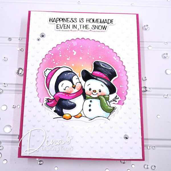 A handmade holiday greeting card featuring two cartoon penguins and a snowman under a pink scalloped circle, with the text 'HAPPINESS IS HOMEMADE EVEN IN THE SNOW' at the top. The card is photographed on a white textured surface with scattered rhinestones.