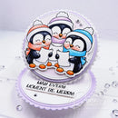 A handmade layered card featuring three penguins in winter clothing singing from paper sheets, with the text 'MAY EVERY MOMENT BE MERRY' on a scalloped white base. The card is decorated with small silver rhinestones and photographed on a white surface.