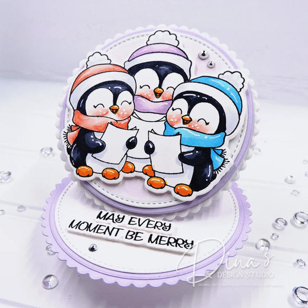 A handmade layered card featuring three penguins in winter clothing singing from paper sheets, with the text 'MAY EVERY MOMENT BE MERRY' on a scalloped white base. The card is decorated with small silver rhinestones and photographed on a white surface.