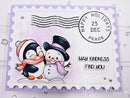 A handmade holiday greeting card styled like a postage stamp, featuring a penguin and snowman illustration, a black 'HAPPY HOLIDAYS 25 DEC PEACE' stamp-like circle, and the words 'MAY KINDNESS FIND YOU.' The card has a lavender scalloped border and is photographed on a white surface with small clear rhinestones around it.