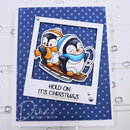 A handmade Christmas card featuring two cartoon penguins sledding on a blue polka-dot background. The card has a white border with the text 'HOLD ON IT'S CHRISTMAS' and is displayed on a white surface with small silver rhinestones.