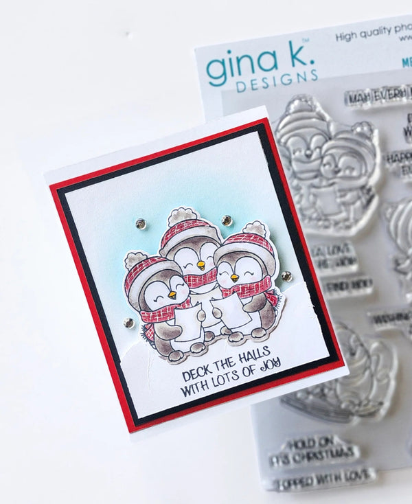 A handmade Christmas card featuring three penguins wearing red and white striped scarves and hats, with the text 'DECK THE HALLS WITH LOTS OF JOY'. The card is shown next to a sheet of Gina K.
